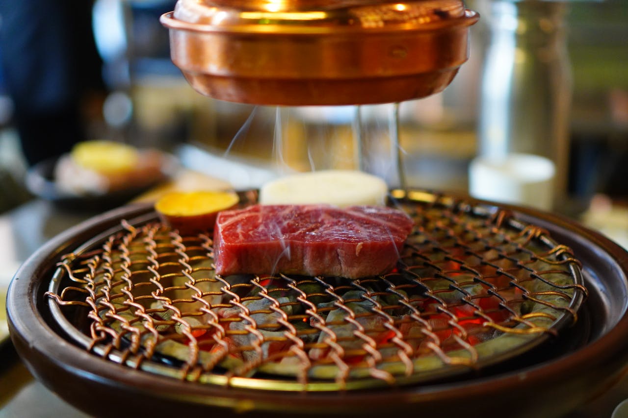 home-img Close-up of raw beef on a Korean BBQ grill, sizzling under a copper hood in a restaurant setting.